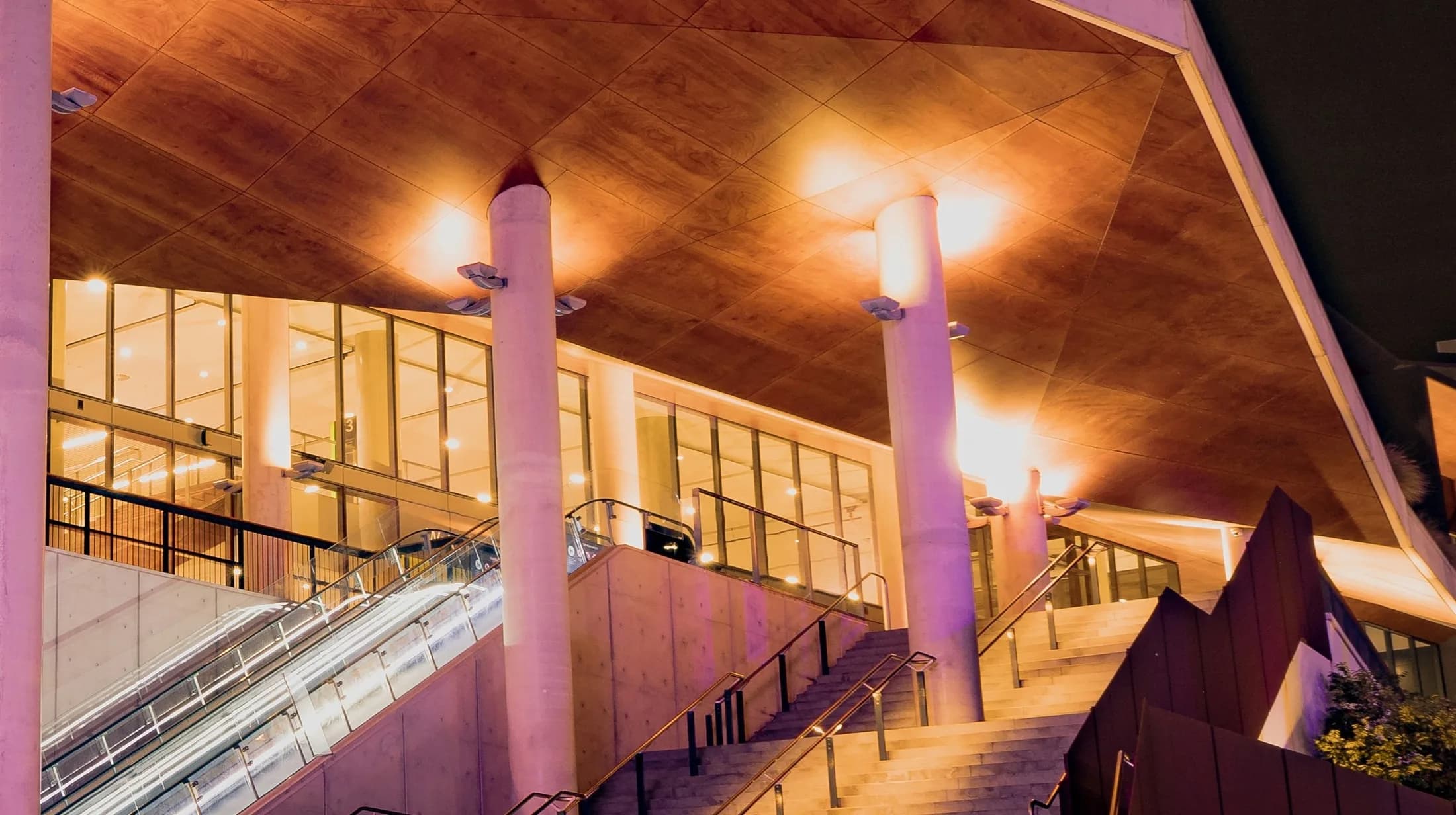A venue entrance at night: escalators and stairs rising under a glowing timber roof between lit columns
